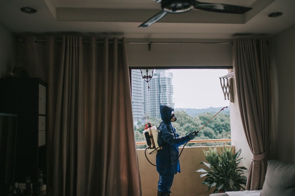 Person in protective gear spraying pesticide from a balcony, overlooking a green landscape and urban buildings, illustrating pest control services.